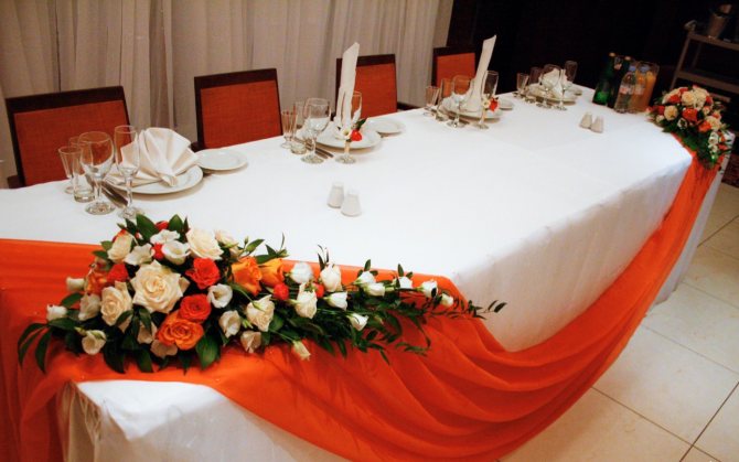 Драпировка стола скатертью и полосой ярко красной ткани, декорированной композицией цветов.
